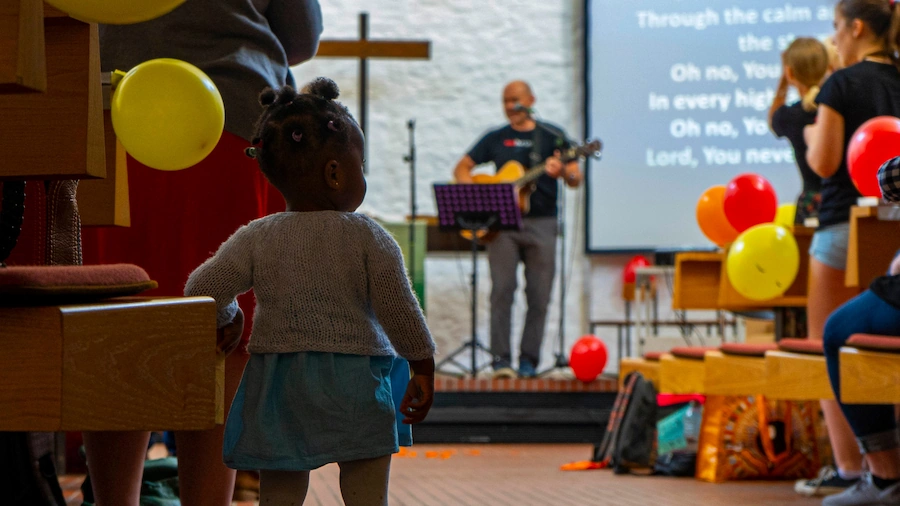 A toddler listening to worship music with a guitar player and colourful balloons in the background.